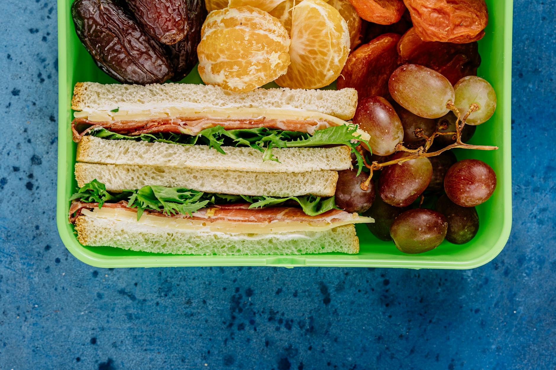 Kolorowy lunchbox z kanapką, winogronami, pomarańczą i daktylami na tle